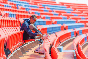 Sport man athlete with a prosthesis on his leg sit on chair of amphitheater to tie shoelaces in the...