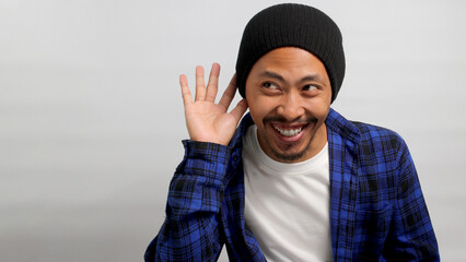 Young Asian man, dressed in a beanie hat and casual shirt, laughs while listening intently, hand...