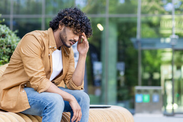 Tired and depressed young indian man sitting on a bench outside, smiling sad and holding his head