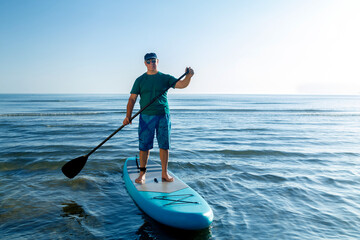 A man in shorts and a T-shirt stands on a SUP board with a paddle near the sea. - obrazy, fototapety, plakaty