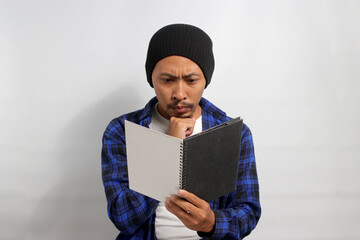 A serious young Asian student, dressed in a beanie hat and casual shirt, is deeply engrossed in...