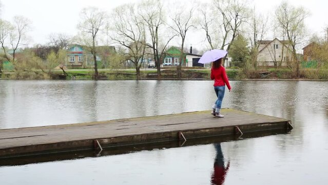 Young woman with purple umbrella goes on wooden pier in rain 