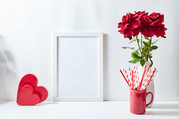 Mockup with a white frame and red roses in a vase, red heart  on a white table. Empty poster frame...