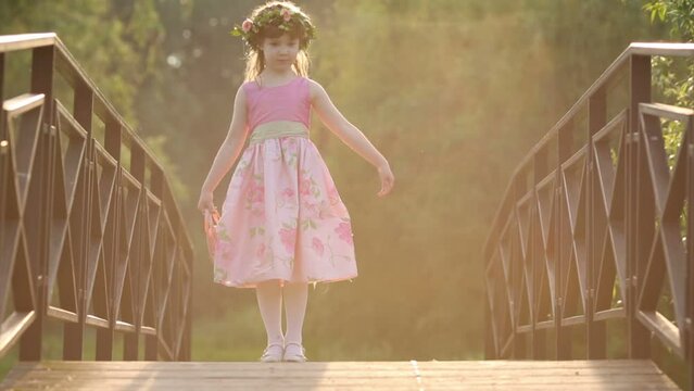 Little girl in pink dress and wreath dances on bridge in park