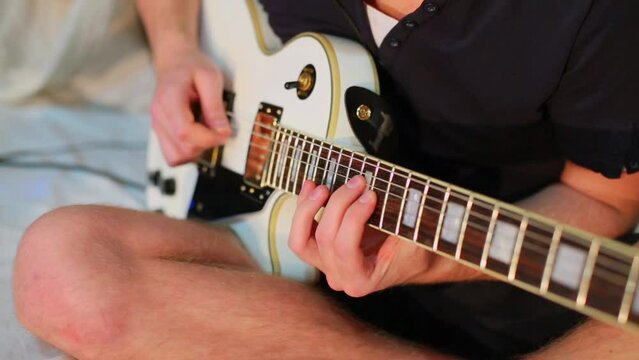 Young musician sits at bed and plays on white guitar