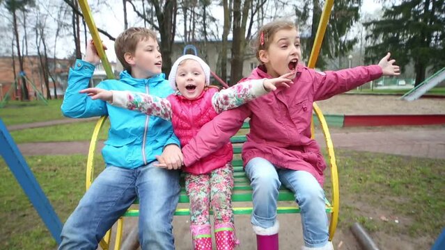 Cheerful boy and two girls in bright jackets shout on swings