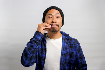 an Asian man, dressed in a casual shirt and wearing a beanie hat, is gesturing for silence by mimicking a zipped mouth, indicating a promise to keep someones secret safe, Isolated on White background