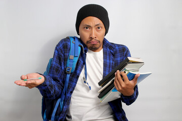 A confused young Asian student, dressed in a beanie hat and casual shirt and carrying a backpack, appears puzzled and shrugs his shoulders while holding books, standing against a white background