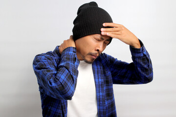 Obraz premium Young Asian man, dressed in a beanie hat and casual shirt, is holding his head, suggesting he's experiencing a headache, isolated on a white background. Healthcare and medical concepts