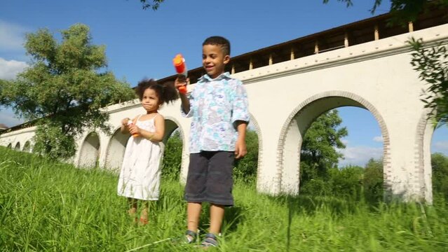 Brother with toy gun and sister stand on grass near aqueduct