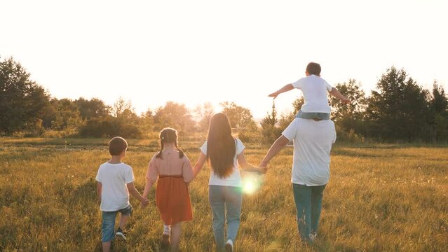 Son sits on father shoulders while family walks with across field at sunset light. Caring parents spend time with children in nature park on vacation. Little children with parents walk across meadow