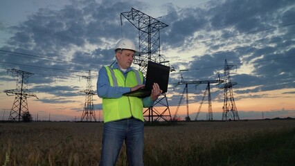 Man worker conducts examination of power transmission lines with laptop in sunset rural field....