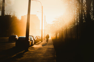 An urban landscape illuminated by the bright morning sun, where a lonely man rides a bicycle to work on a foggy morning. Lifestyle. A shadow. - obrazy, fototapety, plakaty