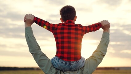 Fototapeta na wymiar Father holds boy on shoulders spreading arms to sides at sunset on field with breathtaking view of nature beauty. Father spends time with little boy. Calm boy feels warmth and care from father