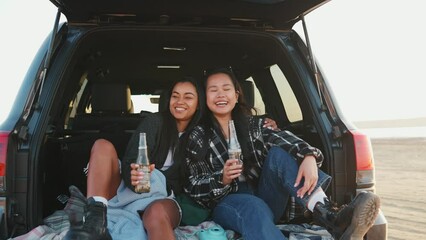 Cheerful multicultural female friends clinking beers and hugging in trunk of car by estuary on sand in summertime