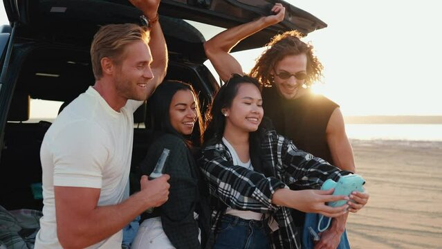 Charming multinational friends take selfie on camera with beer in trunk of car by estuary on sand in summertime