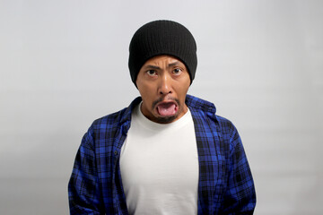 Displeased young Asian man, dressed in a beanie hat and casual shirt, is sticking out his tongue in disgust while standing against a white background