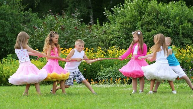 Six happy children pull rope near flowerbeds in summer park