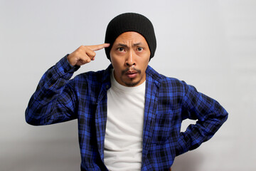 Angry young Asian man in a beanie hat and casual shirt is gesturing with his finger against his...