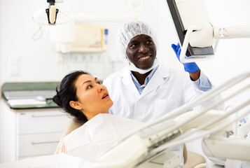 Asian woman sitting on dental chair. African-american man dentist pointing at display.