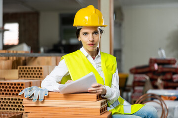 Focused female civil engineer in yellow uniform making notes while controlling workflow and safety on construction site indoors