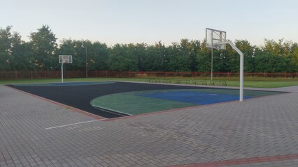 The rubberized basketball court is located among grass lawns and concrete paved areas, next to trees and surrounded by a metal fence. Plastic basketball boards with rings are attached to metal posts