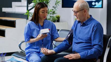 Obraz premium Assistant checking senior man cardiac pulse during cardiology examination in hospital waiting room. Asian nurse measuring patient blood pressure and hypertension using medical tonometer