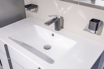 White ceramic sink with chrome faucet in the bathroom. Soap holder and liquid soap dispenser on the wall. Objects have clean lines and modern design