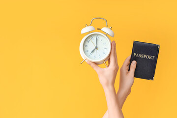 Female hands holding alarm clock and passport on yellow background