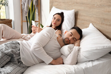 Asleep young man and his wife using mobile phone in bedroom