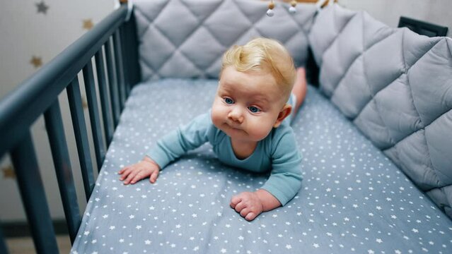 Funny baby boy with adorable plump cheeks lies in his crib. Beautiful blond infant at home.