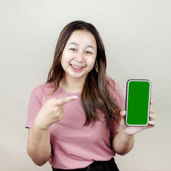 Beautiful smiling Asian woman pointing finger up to empty space while holding her mobile phone on isolated cream background