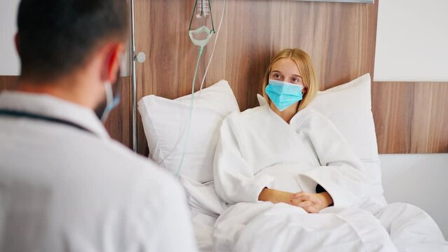 A girl in a hospital room in a medical mask holds her stomach with her hand in pain and listens to doctor with his recommendations