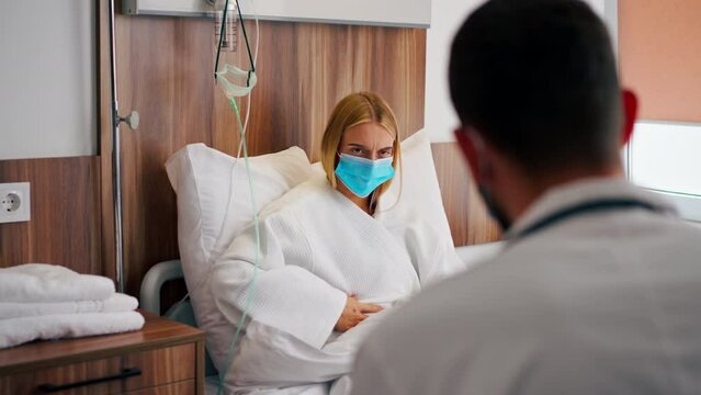 A girl in a hospital room in a medical mask holds her stomach with her hand in pain and listens to doctor with his recommendations