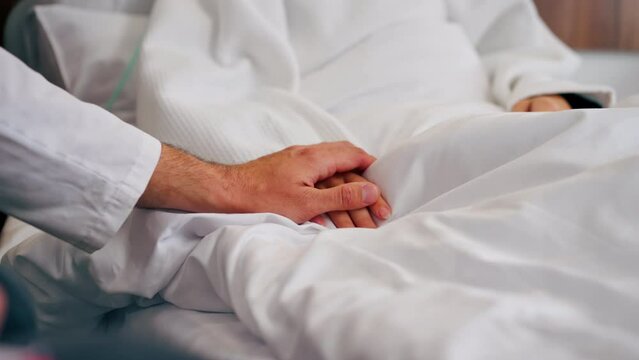 Close-up shot of a male doctor's hand holding a female patient's hand on a bed in hospital ward for moral support