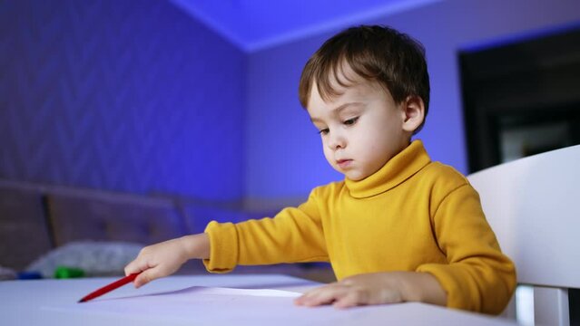 Serious toddler boy sitting at desk drawing with felt-pen. Infant child puts the pen and stands up.