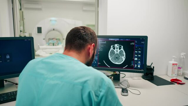A radiologist sits at a table behind a computer monitor and examines a magnetic resonance imaging image during an examination of patient