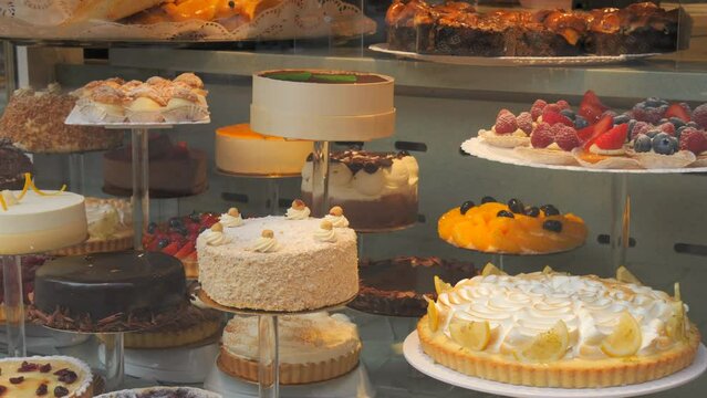 Many different delicious fresh cakes on the counter of the shop, people passing by, reflected in the glass of the window