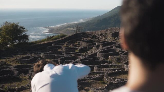 Sunlit summer eve on Santa Tecla, A Guarda. Two young men, one absorbed in the view, the other setting up a camera on a tripod. Ancient Castro, lush hills and Atlantic ocean provide a serene backdrop.