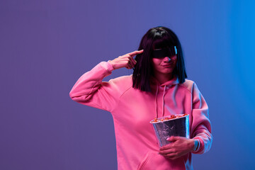Funny confused brunet woman in pink hoodie trendy specular sunglasses with popcorn hold finger on temple posing isolated in blue violet color light background. Neon party Cinema concept. Copy space