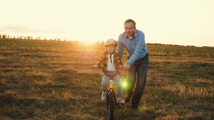 Attentive dad nudges delighted son sitting on bike conveying sense of protection. Papa instils love of physical activity in little boy. Father holds transportation maximizing safety of kid at sunset