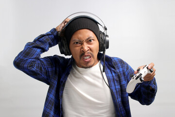 An angry and displeased young Asian man, dressed in a beanie hat and casual shirt, appears stressed while playing a video game using a gamepad on a game console, isolated on white background - obrazy, fototapety, plakaty