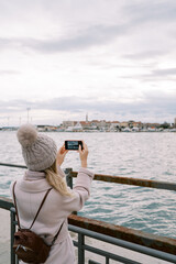 Girl with a backpack stands on the pier at the railing and takes pictures of houses on the seashore with a smartphone. Back view