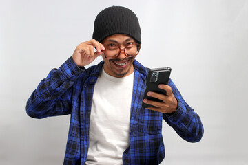 Asian man, dressed in a beanie hat and casual shirt, adjusts his eyeglasses and peeks above them, smiling as he looks at the camera with a mobile phone in hand, while standing against white background
