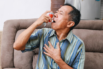 Old man sitting on couch at home holding chest having difficulties breathing and using asthma inhaler to prevent shortness of breath - obrazy, fototapety, plakaty