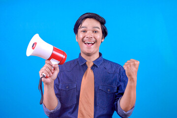 Excited young businessman clenching fist showing victory gesture while holding megaphone on blue...