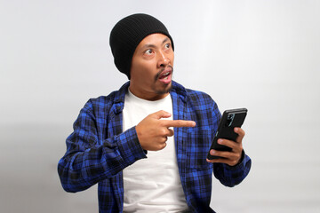 An excited young Asian man, dressed in a beanie hat and casual shirt, points at his mobile phone while standing against a white background