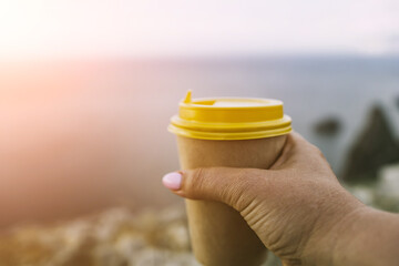 Hand holding Yellow cup with lid, coffee against a backdrop of a blue sky and sea. Illustrating cup and beverage