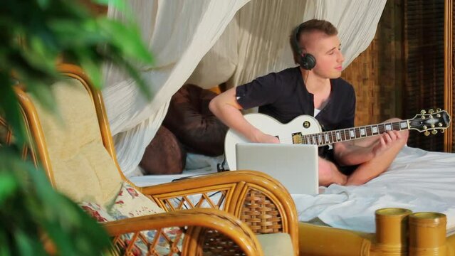 Musician sits at bed with laptop and plays music on guitar