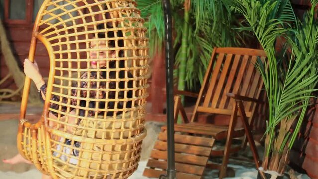 young woman circling in wicker hanging chair near beach house